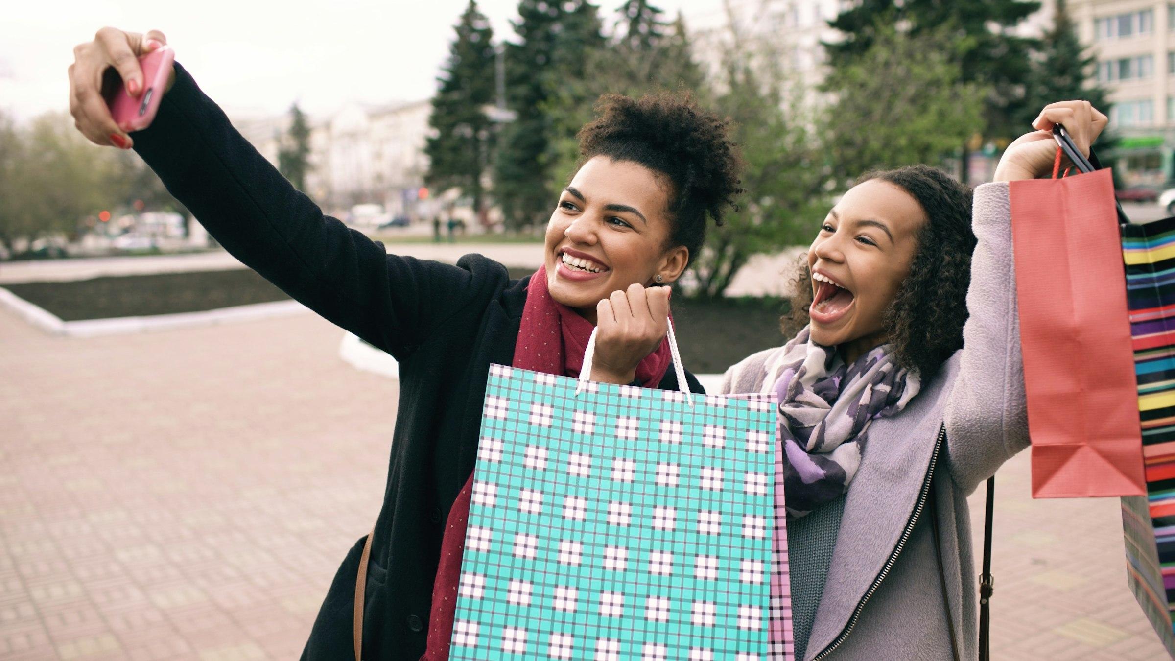 Two friends laughing and taking a selfie with colorful shopping bags